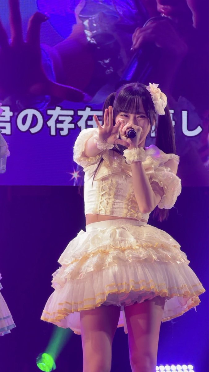 A young idol performs on stage in a frilly cream-colored dress with a large bow, singing into a microphone with one hand raised, while Japanese text appears on the screen behind her. The post's hashtag "#くるみとゆびきり" (Kurumi to Yubikiri) suggests this is a performance by or featuring the idol Kurumi, matching the caption's description of her as "divine" and radiant on stage.