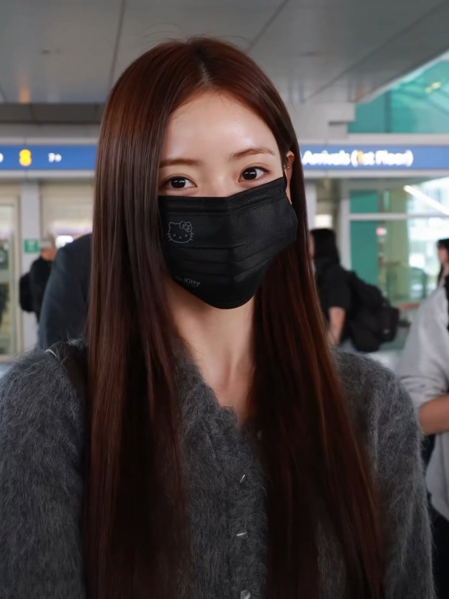 A young woman with long, sleek brown hair stands at what appears to be an airport, wearing a black face mask adorned with a small Hello Kitty design and a cozy gray cardigan. Behind her, an "Arrivals (Exit)" sign is partially visible, suggesting the caption "Kawaii" refers to the cute Hello Kitty detail on her mask.