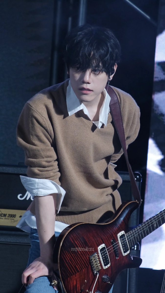 A young man with dark hair wearing a brown sweater over a white collared shirt, holding a red electric guitar, with a watermark 'mylmdru' visible.