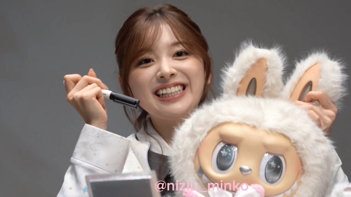 A smiling young woman with brown hair holds a marker and poses next to a fluffy white plush toy with bunny ears, with the watermark '@nizu_minkoi' visible.