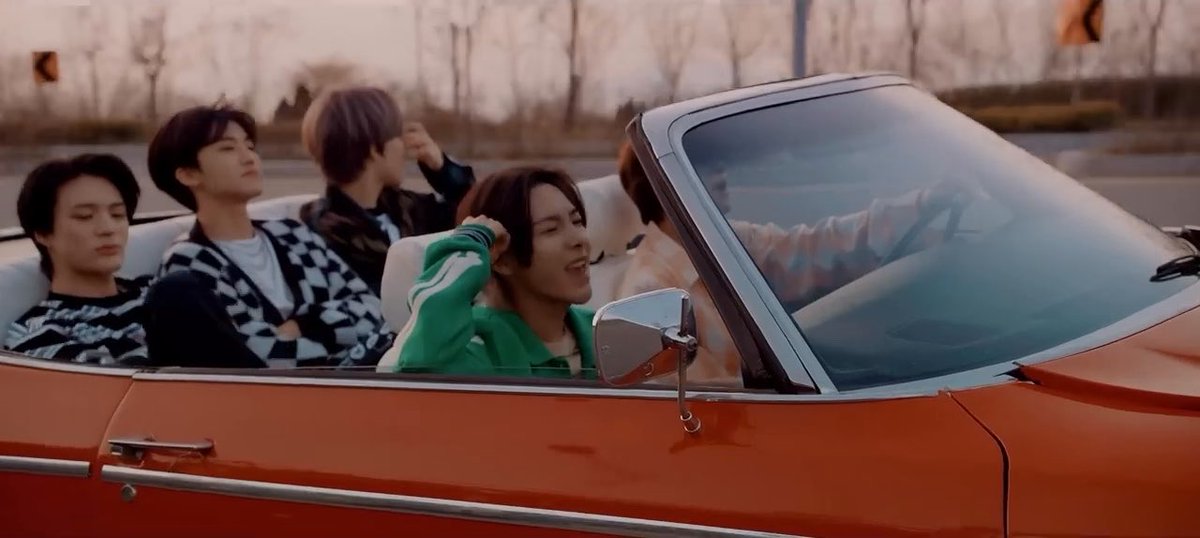 Five young men ride in a red convertible car, with the driver wearing a green jacket.