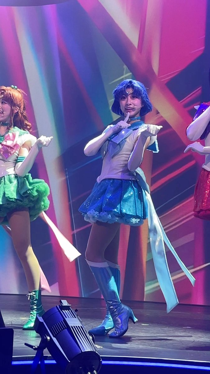 A performer dressed as Sailor Mercury from Sailor Moon in a blue and white sailor-style costume with a blue wig, posing on stage alongside another performer in a green outfit.