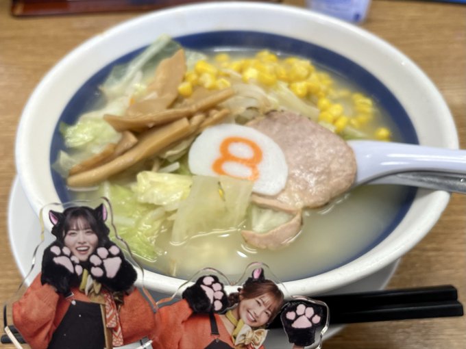 A bowl of ramen with corn, vegetables, and a narutomaki fish cake decorated to look like a cat, accompanied by two acrylic standees of smiling Japanese idols with paw decorations.