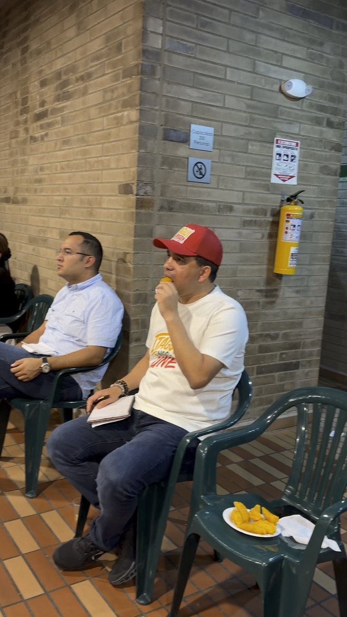 Two men sit in green plastic chairs in a brick-walled room; one wears a red cap and white t-shirt while eating, with a plate of fried food on the chair beside him.