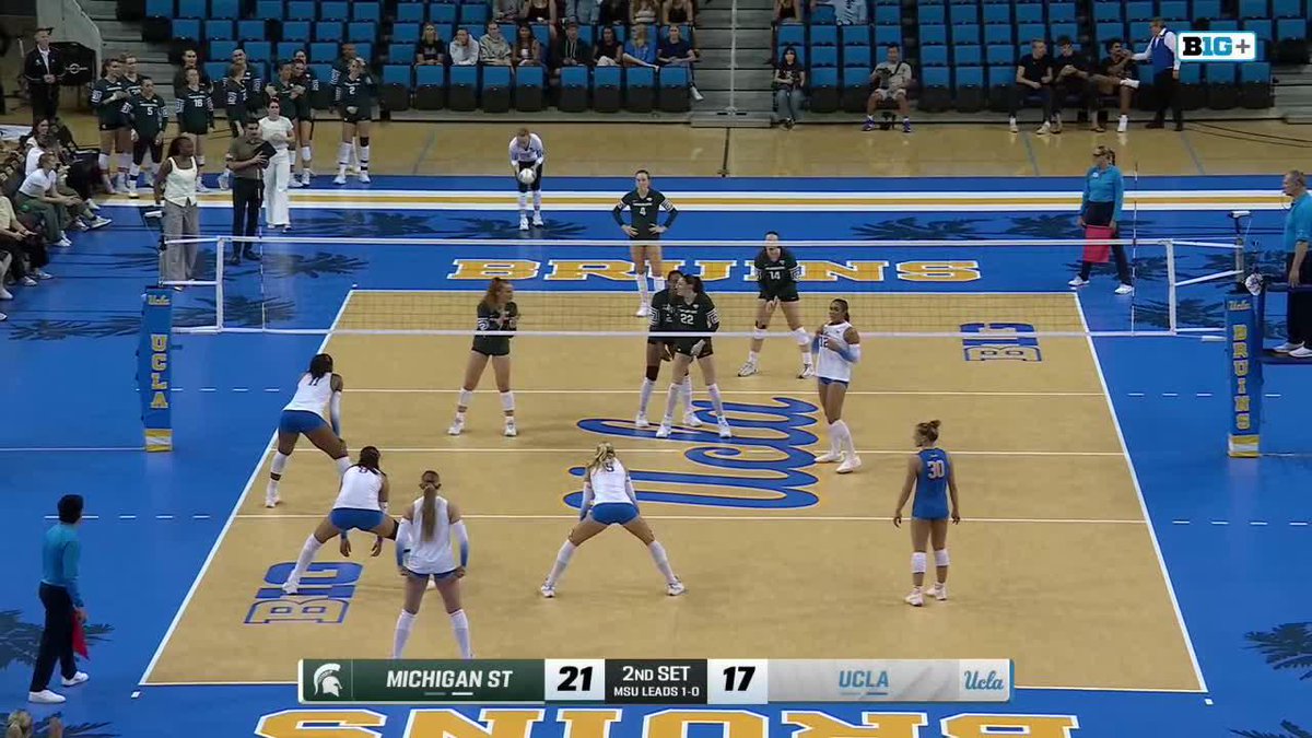 A women's college volleyball match between Michigan State (green jerseys) and UCLA (white/blue jerseys), with the scoreboard showing "MICHIGAN ST 21, 2ND SET 17, UCLA" at UCLA's court.