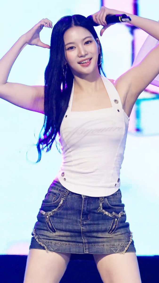 A young woman with long dark hair poses with her arms up, wearing a white halter top and denim skirt on a stage with purple lighting.