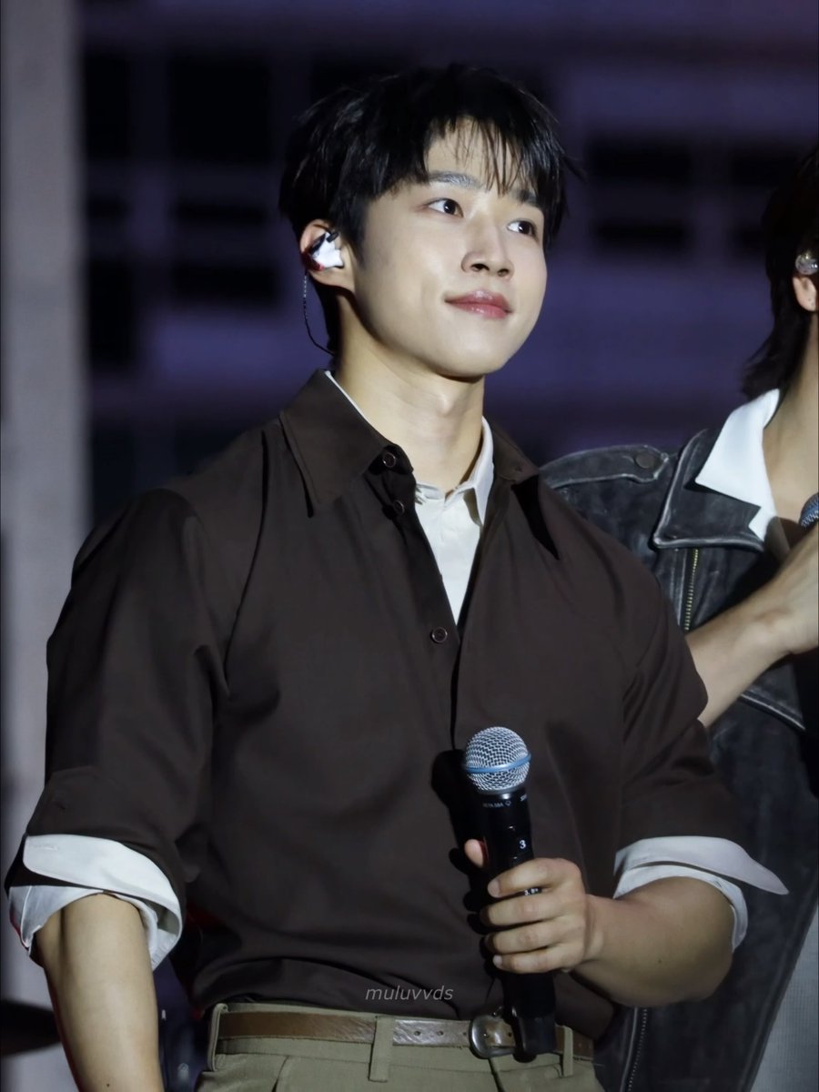 Photograph of a young male performer wearing a brown shirt and in-ear monitors while holding a microphone, with a watermark reading 'muluvvds'.