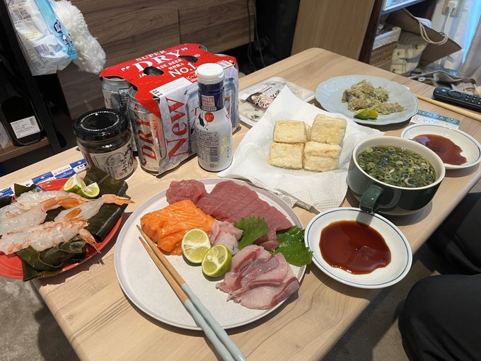 A Japanese meal spread on a wooden table includes a plate of sashimi (salmon, tuna, squid), shrimp, fried tofu, miso soup, dipping sauce, and beer cans.