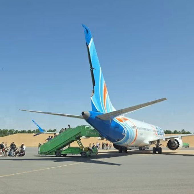 A FlyDubai aircraft with blue and orange livery sits on a tarmac with mobile boarding stairs attached, while passengers board under a clear blue sky. The image accompanies reporting about FlyDubai co-pilot Hamam al-Hammami, an Omani national allegedly banned from flying by Oman due to radical views, who is accused of attempting to crash a jet full of Israeli passengers.