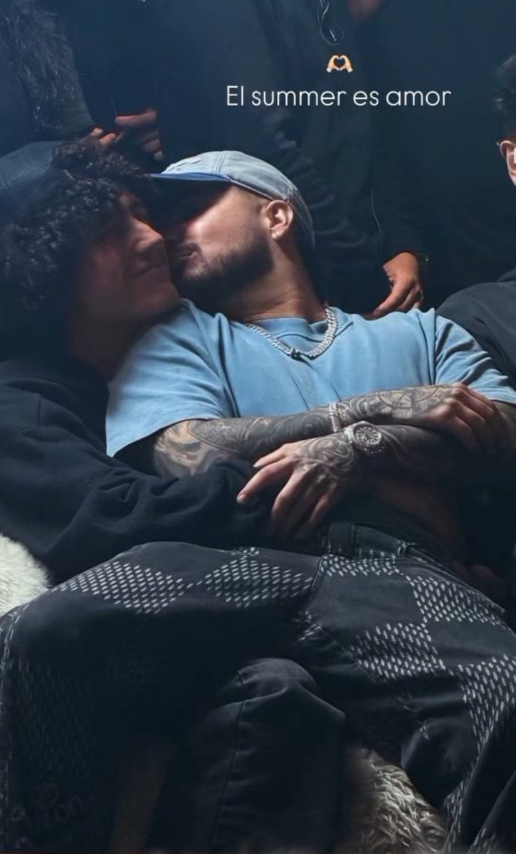 Two men sit close together with their faces nearly touching; one wears a blue cap and shirt with tattoos on his arms, and the Spanish text 'El summer es amor' appears at the top.