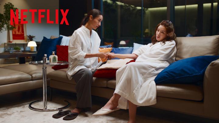 The Netflix logo appears in the top left corner of an image showing two women in white robes sitting on a couch in a modern living room, with one handing the other a package—likely a promotional scene from a Japanese drama. The caption suggests this is a teaser for an upcoming Netflix release, with fans eagerly anticipating additional official videos featuring characters Rin Rinri and Numata Fumi.