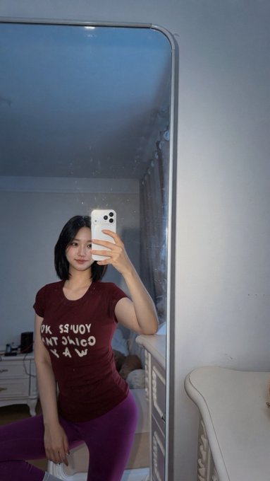 A woman takes a mirror selfie with her phone in a bedroom, wearing a maroon t-shirt with white text and purple leggings.