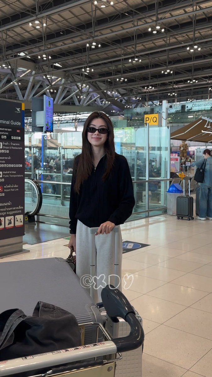 A stylish woman with long dark hair and sunglasses stands confidently at an airport terminal, posing with her luggage trolley near a "Terminal Guide" sign indicating shops, restaurants, and the Airport Rail Link, with "P/Q" and "Observatio[n]" signage visible in the background. The caption "So handsome, a Lena" celebrates her chic black blouse and sunglasses look, fitting the glamorous travel aesthetic suggested by the hashtags referencing Chanel Spring Summer and Paris Fashion Week.