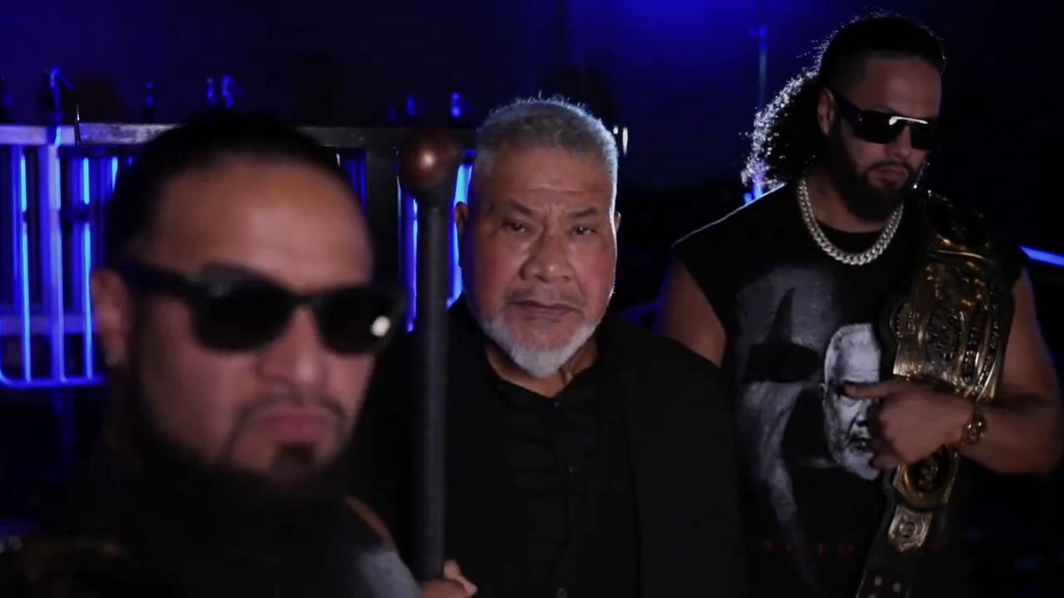 Three men stand together in a dimly lit arena setting with blue lighting, conveying a serious and intimidating demeanor. The man on the left wears dark sunglasses, the central figure with gray hair and a goatee holds a staff, and the man on the right sports sunglasses, a chain necklace, and a championship belt over a sleeveless shirt. The image visually reinforces the post's caption about WWE Tag Team Champions The MFTs issuing a warning to Bakusai.