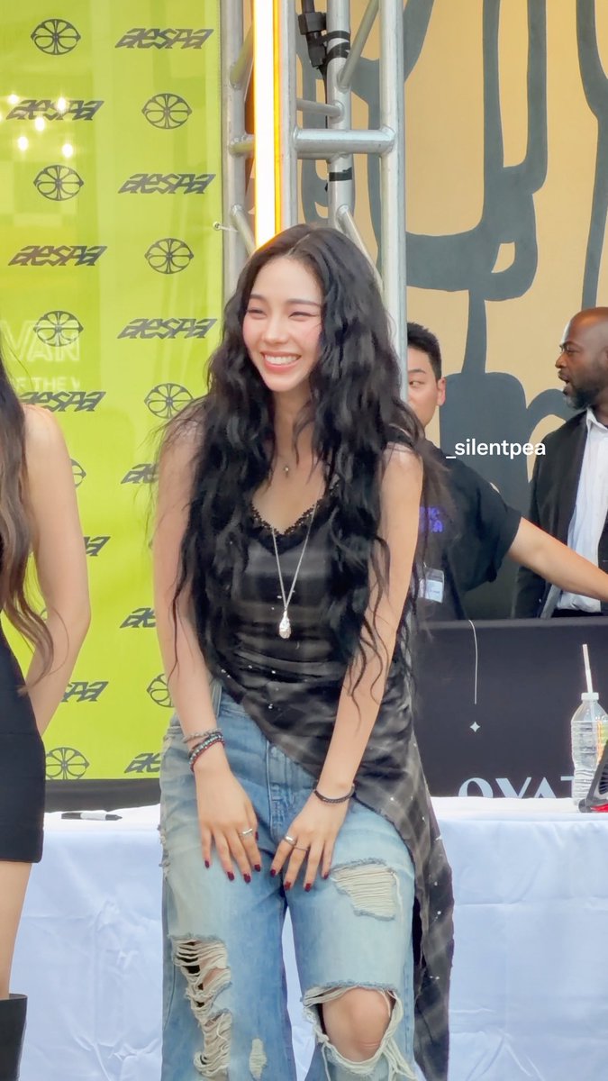 A smiling young woman with long dark wavy hair wearing a black tank top and ripped jeans, standing in front of a backdrop with 'AESPA' text, watermarked '_silentpea'.