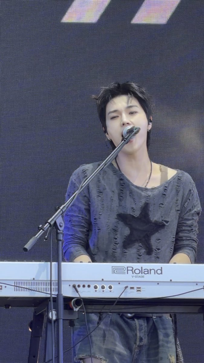 A young man with dark hair plays a Roland V-Stage keyboard while singing into a microphone, wearing a distressed grey long-sleeved shirt.