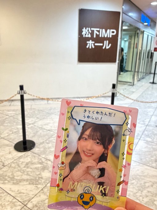 A hand holds up a pink photo card of a young woman named Mizuki in front of the entrance to the 松下IMPホール (Matsushita IMP Hall) in Japan, with a speech bubble reading "きてくれたんだ！うれしい！" ("You came! I'm so happy!").