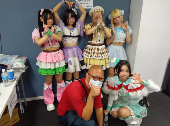 A group photo taken backstage shows four young women in elaborate, colorful idol costumes posing playfully, while a man in a red shirt and mask and a woman in a green and red outfit kneel and pose in the foreground.