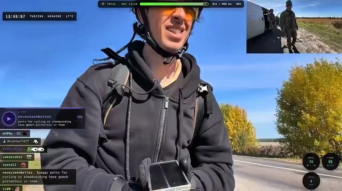 A cyclist wearing a helmet, sunglasses, and a black hoodie livestreams from Turiisk, Ukraine, at 13:08:57 with 915 of 968 km completed (95%) on Day 13/15, while a picture-in-picture view in the corner shows him being confronted by military personnel beside a van on a rural road.