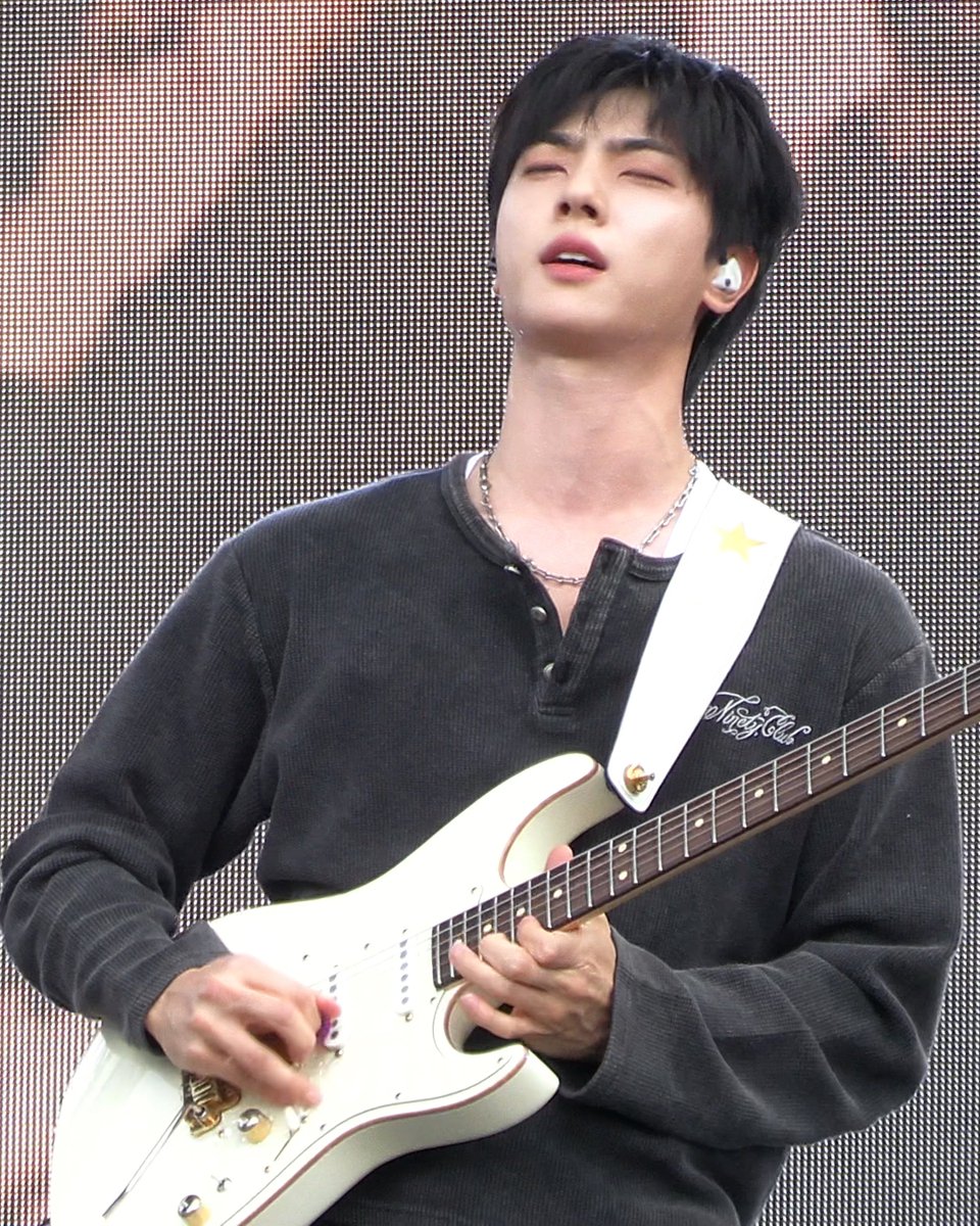 A male guitarist with dark hair, wearing a dark gray henley shirt and a silver chain necklace, plays a white electric guitar on stage with his eyes closed in concentration.