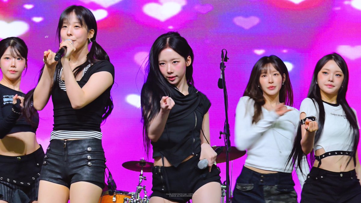 A five-member girl group performing on stage in black and white tops, with the lead singer holding a microphone against a pink background with heart shapes.