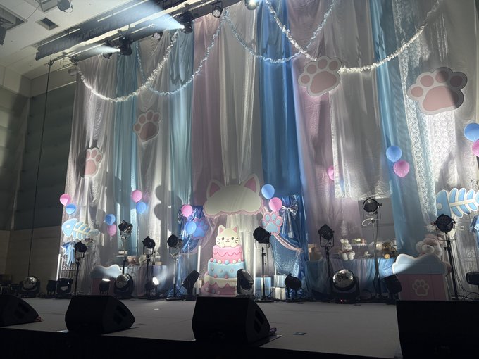 A stage decorated with pastel blue and pink curtains features paw print cutouts, a maneki-neko cat cutout, a fish skeleton design, and various balloons and lights.