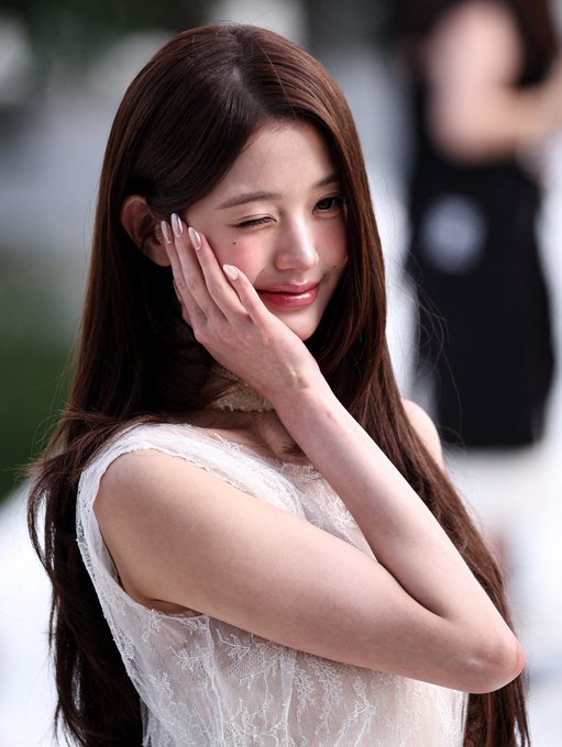 A photograph of a smiling young woman with long brown hair in a white lace top, winking and touching her face.