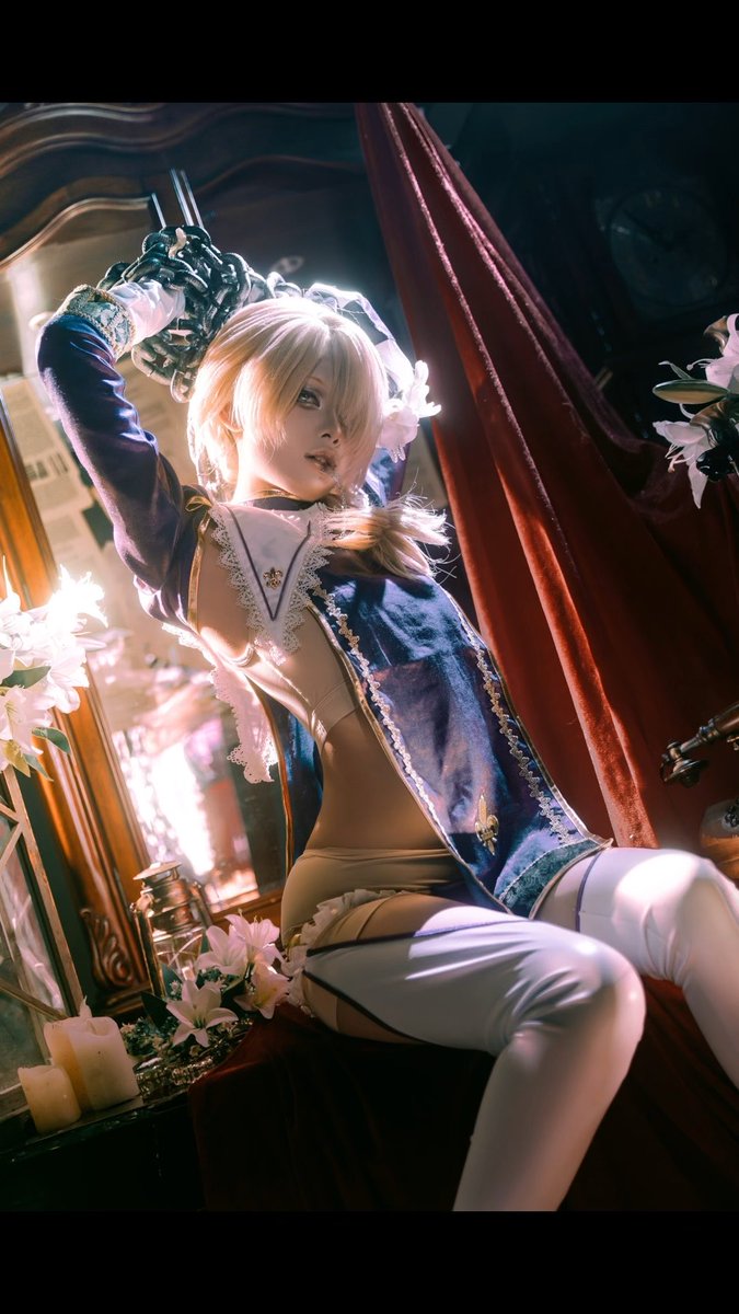 A cosplay photo of a blonde woman in a blue and white outfit with white thigh-high stockings, posed in an ornate setting with curtains and flowers.
