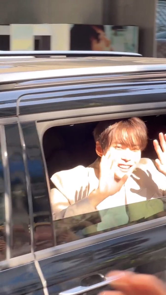 Doyoung from NCT waves enthusiastically from inside a vehicle, smiling brightly through the open window as he greets fans, perfectly capturing a candid fan interaction moment.