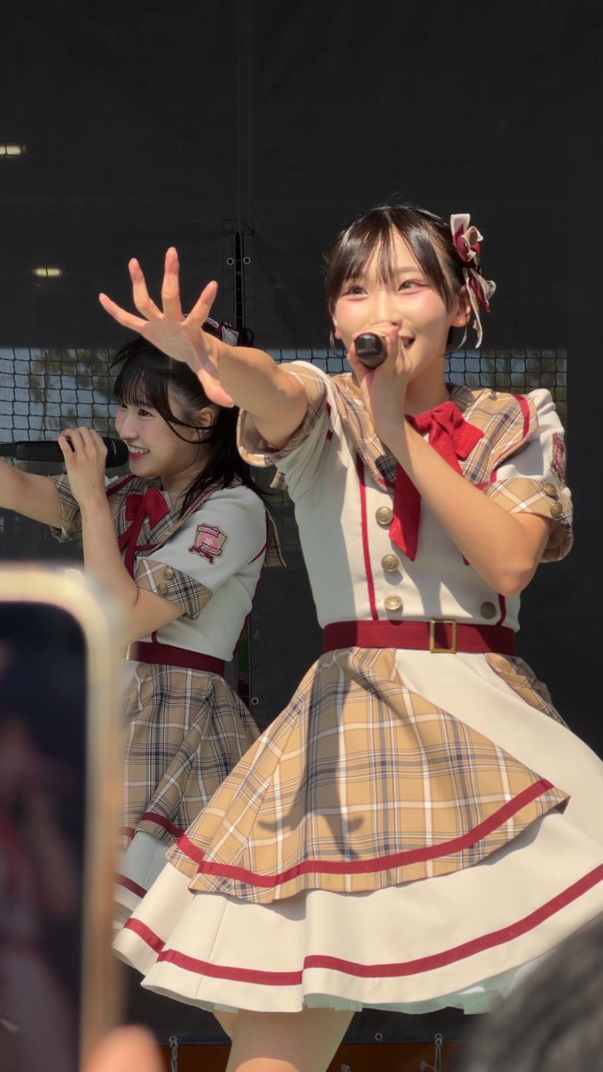 Two young women in matching beige plaid idol costumes with red bows perform on stage, both holding microphones and reaching out toward the audience.