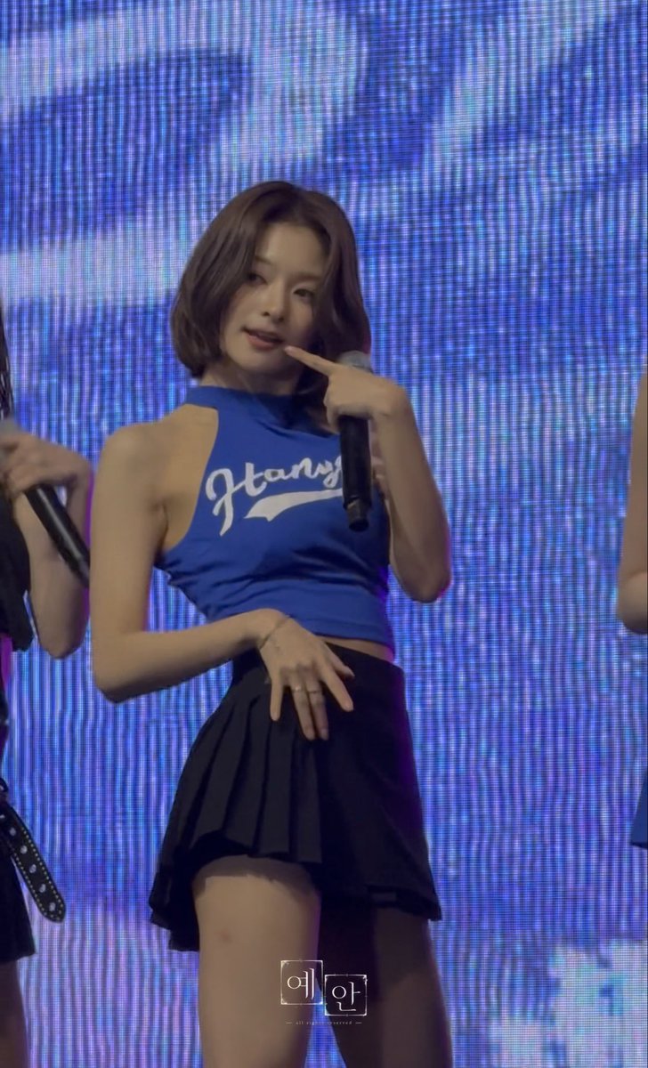 A woman with short brown hair performs on stage in a blue 'Hangang' cropped tank top and black pleated skirt, pointing at her face while holding a microphone against a blue lit backdrop.
