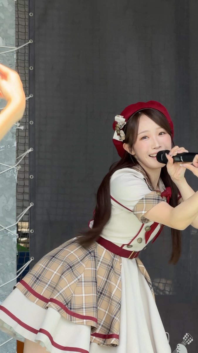 A smiling young woman with long brown hair wearing a red beret with a bow and a plaid/cream dress with red trim, holding a microphone as if performing on stage, with a dark mesh background behind her.