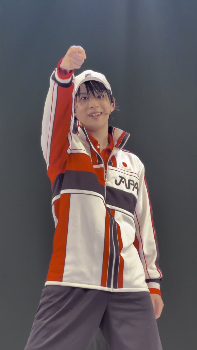 A young performer on stage wearing a red, white, and navy blue 'JAPAN' tracksuit jacket and a FILA cap, smiling and pointing confidently at the audience.