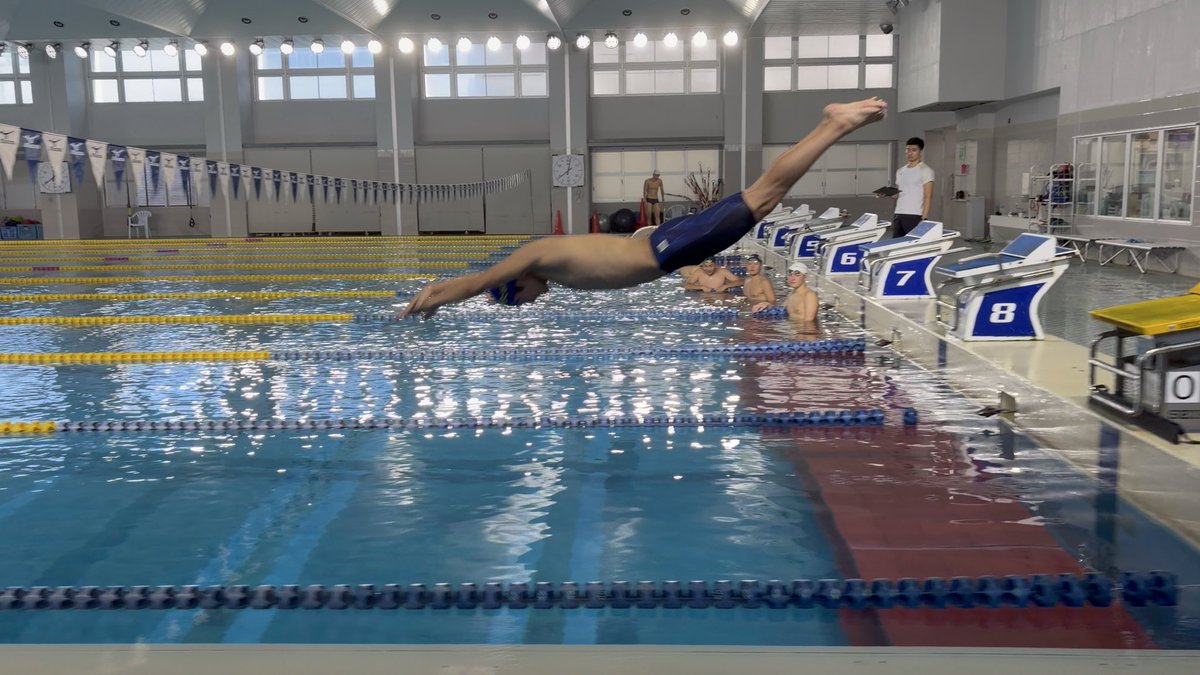 A swimmer in blue trunks dives mid-air into a competition pool, with lane numbers 6, 7, and 8 visible on the starting blocks behind him. The image captures a training moment that reflects the swimmer's dedication to improvement, aligning with the post's goal of breaking through to the Japan Championships.