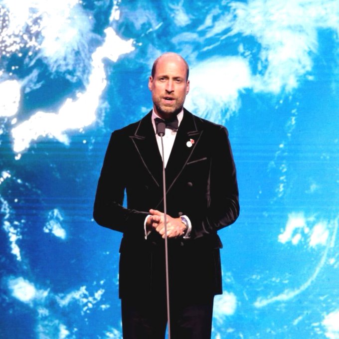 Prince William, dressed in a black tuxedo with a bow tie, stands at a microphone giving a speech against a large blue backdrop displaying an image of Earth from space.
