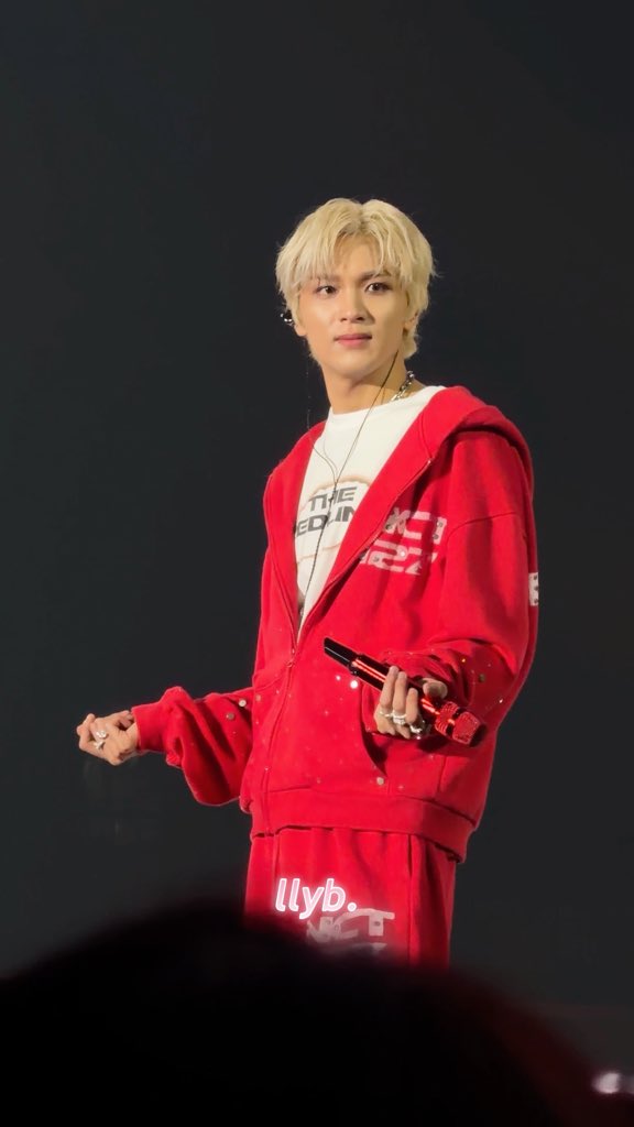 A young man with platinum blonde hair stands on stage in a matching red tracksuit, holding a microphone while looking toward the camera. His hoodie and t-shirt feature visible text including "THE" and "WORLD," along with "llyb" printed on his shorts, capturing a relaxed concert moment that aligns with the casual vibe of "beneran di goyang" (really shaking/grooving).