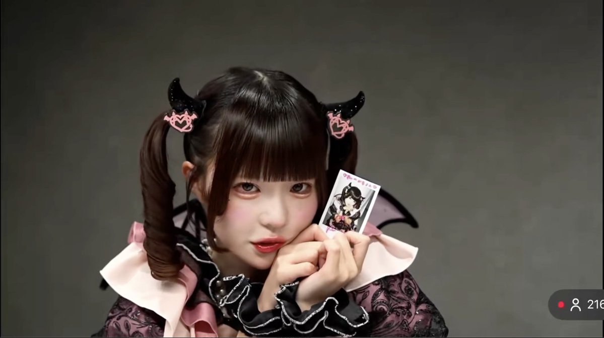 A person in gothic-style cosplay with pink ribbons and dark twin-tails holds up a small photo card against a gray backdrop, with '216' visible in the corner.