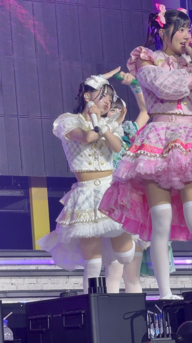 Performers in elaborate pink and white frilly idol costumes perform on a stage, with one member in white holding a microphone and making a playful expression.