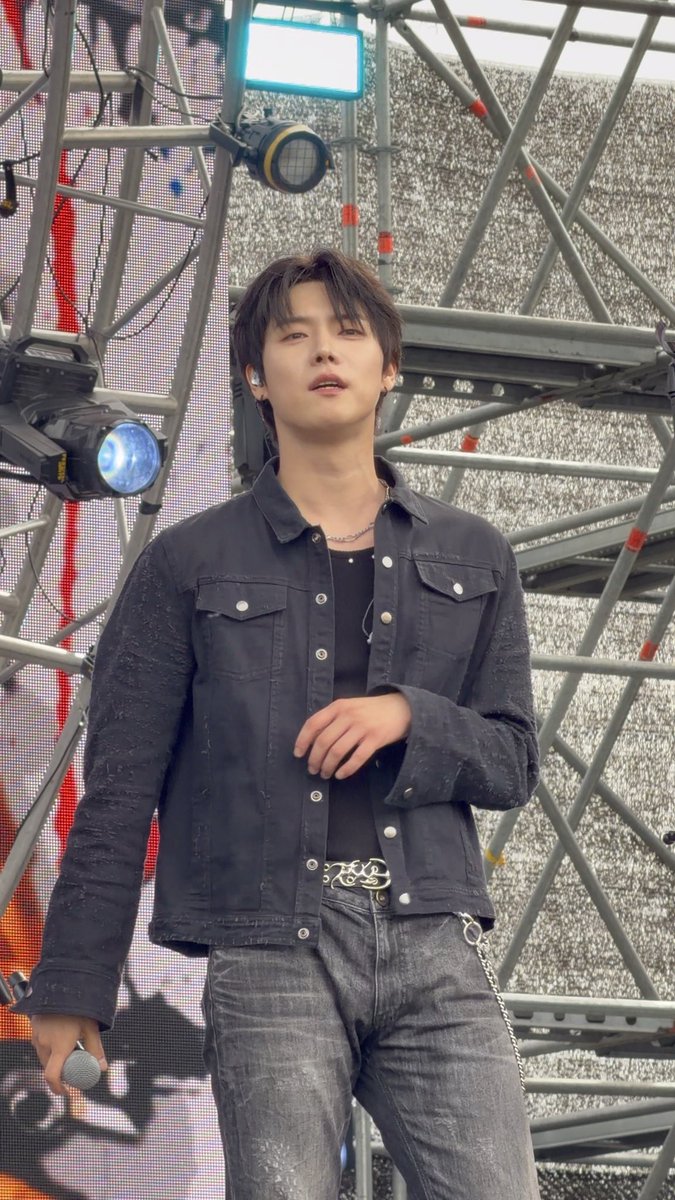 A young male performer stands on stage in a black denim jacket and dark jeans, holding a microphone, with stage lighting and metal scaffolding visible behind him.