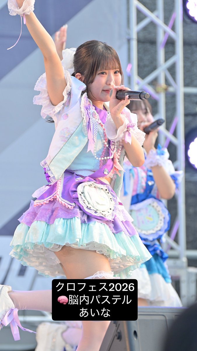 A young female idol performs on stage in a colorful pastel purple and blue frilly outfit, holding a microphone, with Japanese text overlay reading 'クロフェス2026 🧠脳内パステル あいな'.