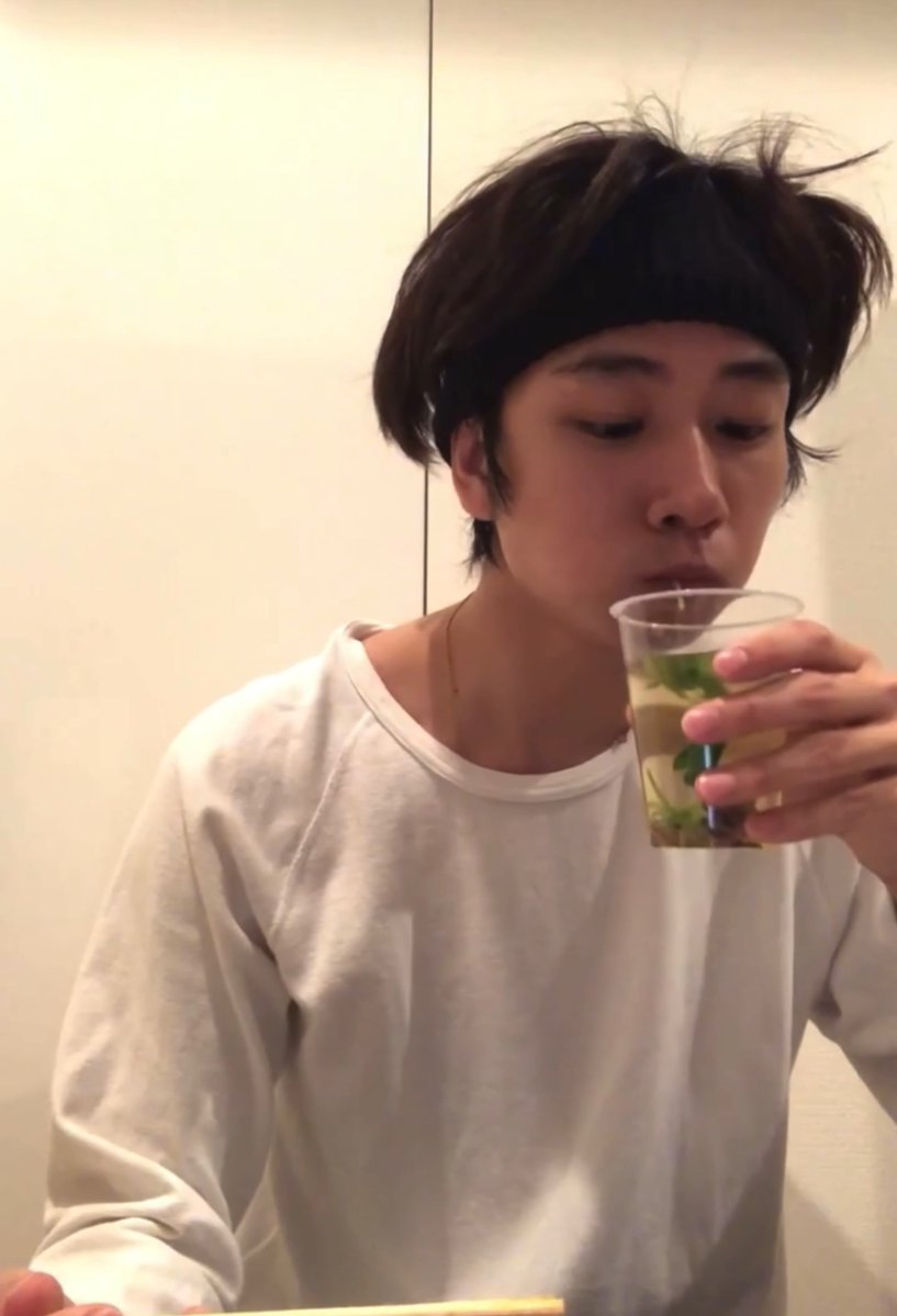 A young man in a white shirt drinks from a clear plastic cup containing water and small green plants, in what appears to be a casual indoor setting with plain white walls behind him. The image humorously ties to the aquarium caption, as he is sipping from a cup that resembles a makeshift fish habitat for medaka (Japanese rice fish).