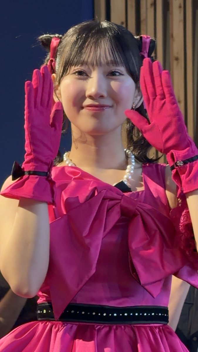 A smiling young woman wearing a bright pink dress with a large bow, matching pink gloves, and pearl necklace, posing with her hands raised near her face at what appears to be an event.