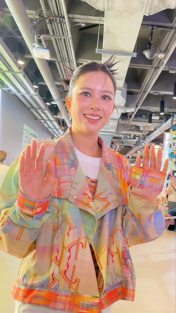 A smiling woman with her hair up poses indoors, holding up her hands, while wearing a vibrantly patterned, colorful jacket over a white shirt.