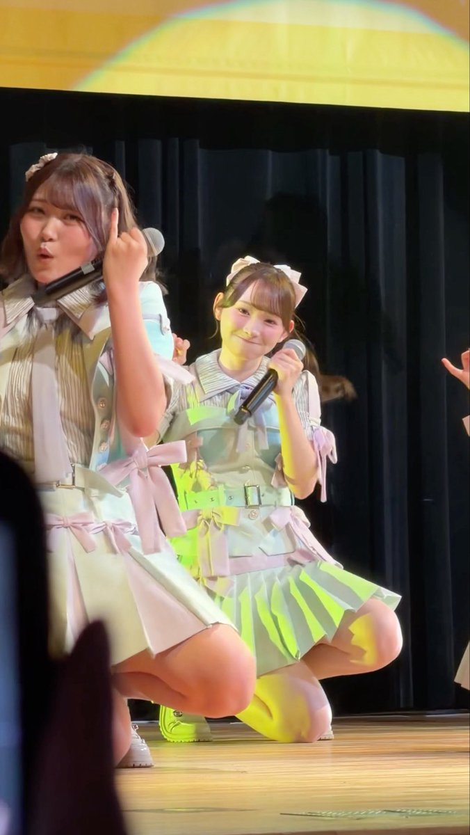 Two young performers in matching pastel idol outfits crouch on stage holding microphones; one makes a middle finger gesture.