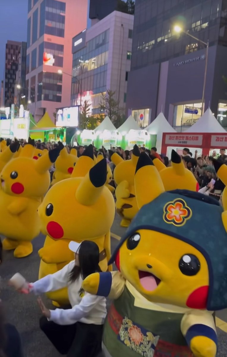 A parade of Pikachu mascots marches through a city street at dusk, with one Pikachu in the foreground wearing a traditional Korean hanbok outfit and a green jeogori-style hat adorned with a flower emblem, while crowds watch near a Land Rover building and booths with Korean signage. The "camouflaged Ditto" reference relates to how Pikachu itself is essentially a Ditto—the Transform Pokémon—disguised in elaborate cultural costumes, showcasing the organizers' attention to detail by dressing the mascots in traditional attire that reflects local heritage.