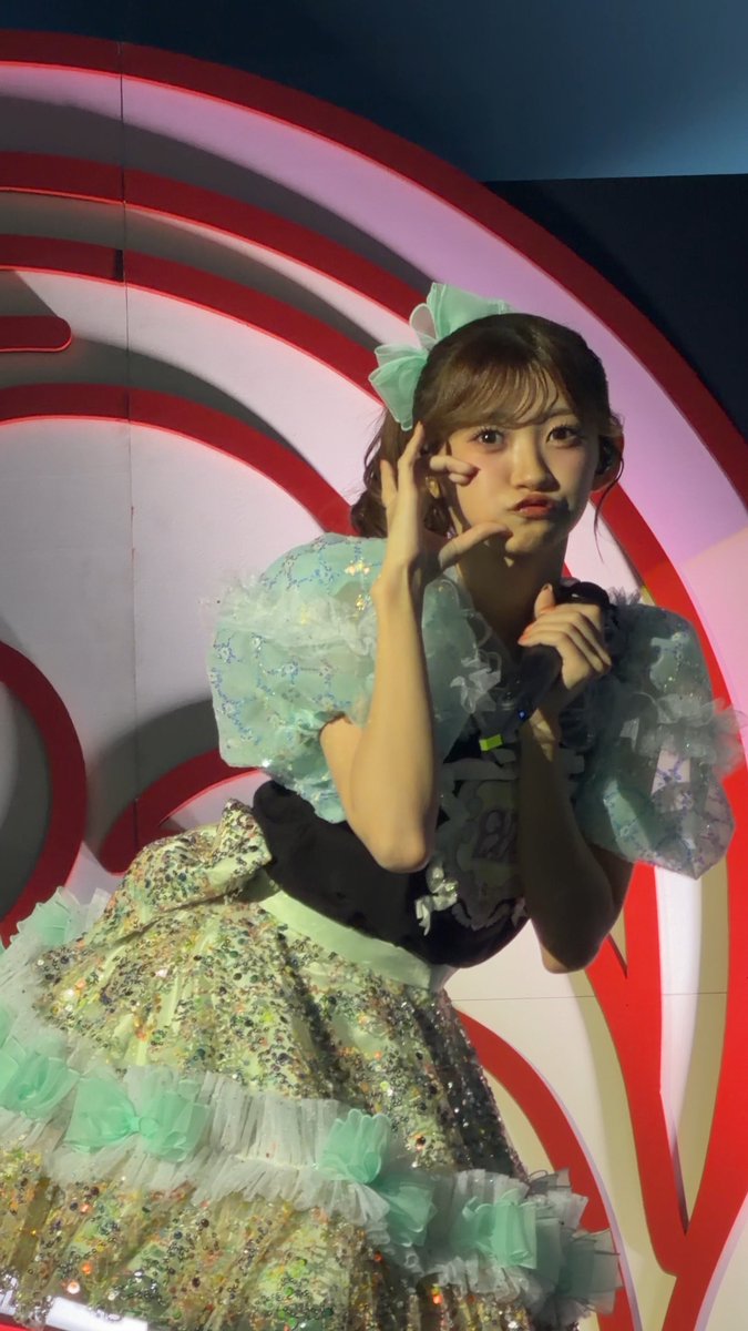 A young performer in a sparkly mint-green and white idol dress with bows and frills, holding a peace sign near her eye against a red and white backdrop.