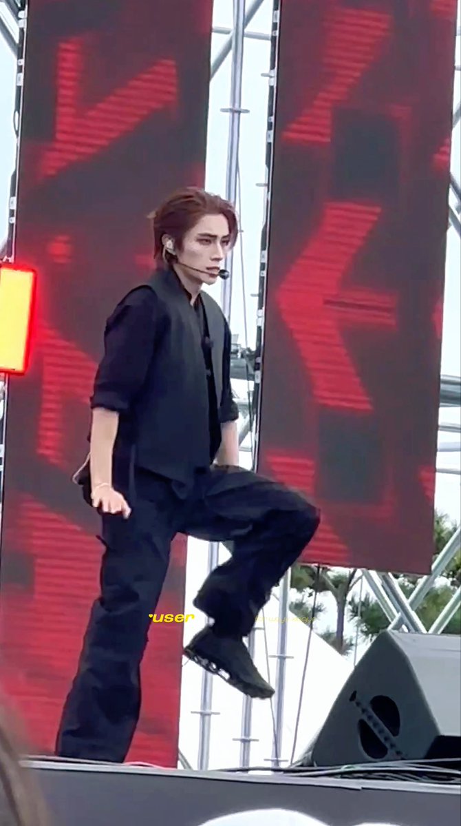 A performer with reddish-brown hair, wearing a black vest and pants over a dark shirt, performs on stage with a headset microphone. The text '*useren' is visible at the bottom.