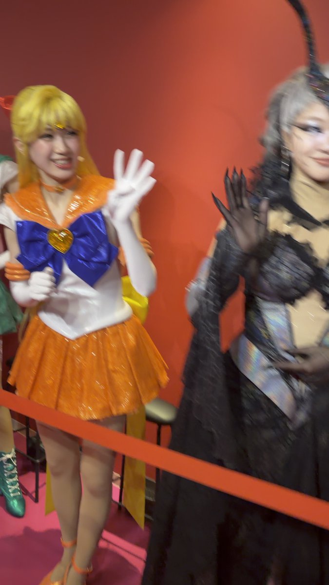 Two women in elaborate costumes wave at the camera; one is dressed as Sailor Venus in an orange and white outfit, while the other wears a dark, horned ensemble.