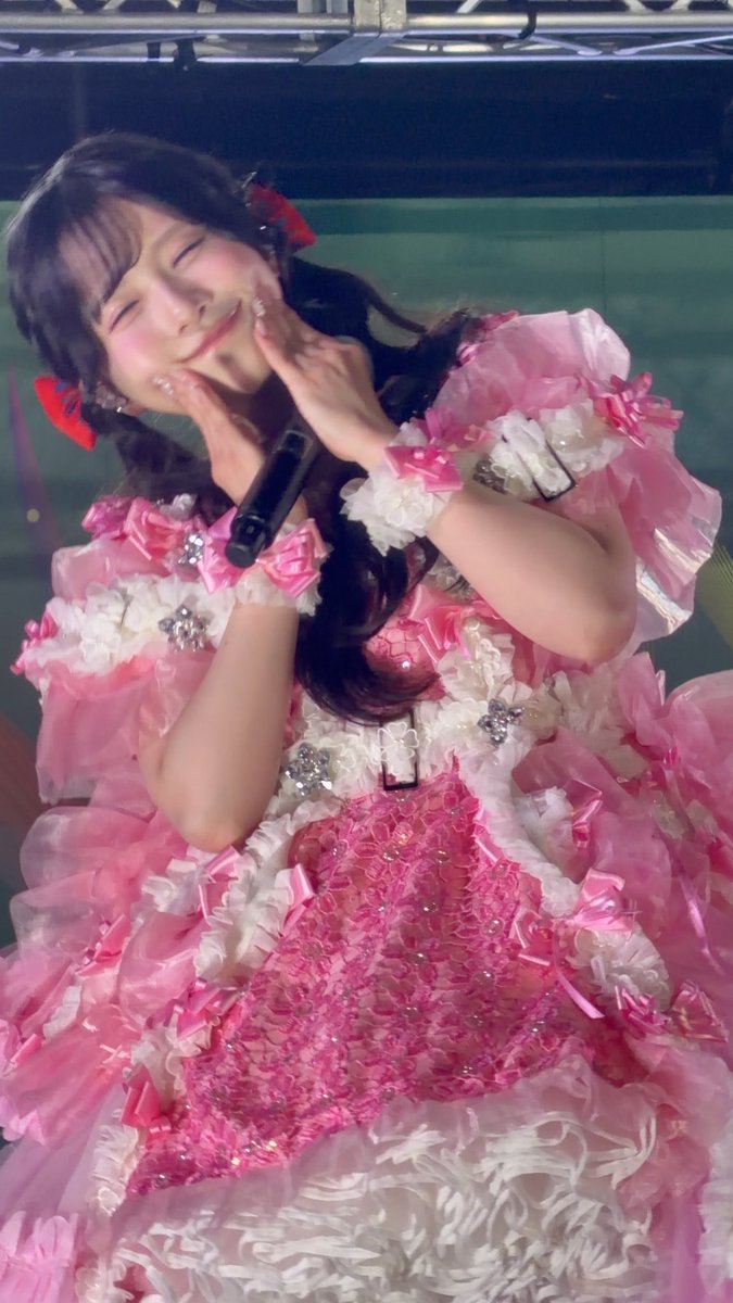 A performer in an elaborate pink frilly dress with rhinestone accents smiles and poses with hands near her face while holding a microphone on stage.