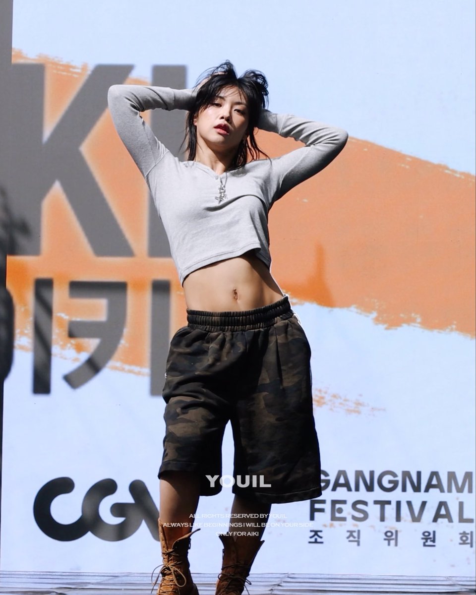 A photo of a woman in a grey crop top and camouflage pants on stage, with text 'YOUIL', 'CG', 'GANGNAM FESTIVAL' and Korean characters visible on the backdrop.